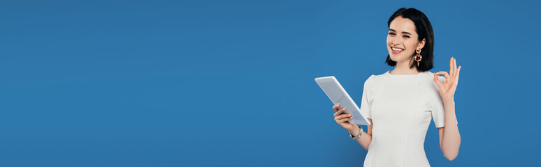 panoramic shot of smiling elegant woman holding digital tablet and showing okay sign isolated on blue