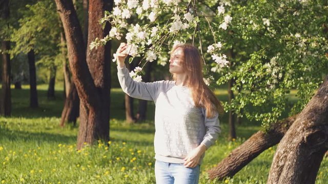 Beautiful caucasian girl on a background of a blossoming tree takes a selfie, straightens her hair