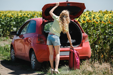 Young girl in front of a red car with a suitcase in her hands.