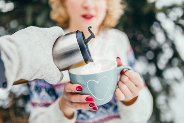 A hand with a thermos pours hot tea or coffee into the cup that a girl is holding in the forest -...