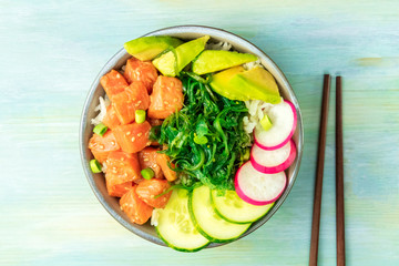 Salmon poke with avocado, wakame and cucumber, shot from the top on a teal blue background with chopsticks