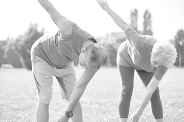 Black and white photo of Active flexible senior couple stretching leg in park on sunny day