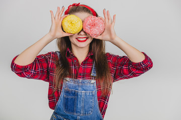 Lovely young female hiding her eyes behind two big yellow and pink doughnuts, lovely smiling.