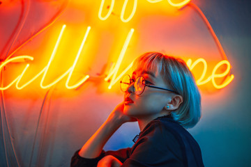 Young adult asian female wearing glasses in front of neon light sign, shallow selective focus