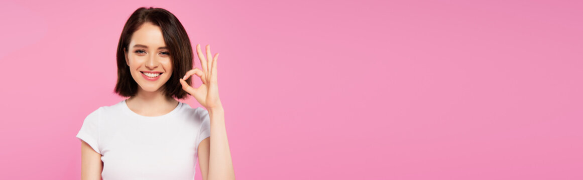 panoramic shot of smiling pretty girl showing okay sign isolated on pink