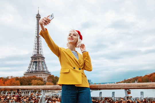 Happy smiling tourist girl makes selfie in Paris with view of Eiffel tower. Lady wearing red beret, yellow blazer. Travel, tourism, autumn vacation, advertising concept. Copy, empty space for text