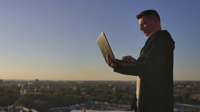Programmer a hacker is on the roof with a laptop at sunset says error code on the keyboard and looking at the city view