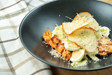 Caesar salad with croutons, parmesan, chicken, egg in a black stylish bowl. Restaurant serving. Selective focus, wooden background