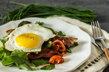 Appetizing breakfast with fried eggs and salad leaves. From above delicious meal with bacon egg tomatoes and green leaves served on white plate over gray textured background
