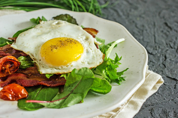 Appetizing breakfast with fried eggs and salad leaves. From above delicious meal with bacon egg tomatoes and green leaves served on white plate over gray textured background