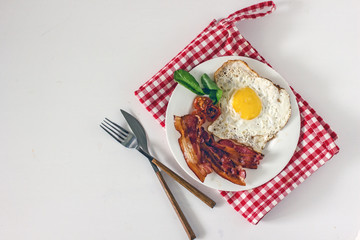 Homemade breakfast with fried eggs and bacon. Top view on a white plate with fried egg and fried bacon, decorated with greens. The plate stands on a napkin in a red and white cage, next to a knife