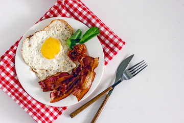 Homemade breakfast with fried eggs and bacon. Top view on a white plate with fried egg and fried bacon, decorated with greens. The plate stands on a napkin in a red and white cage, next to a knife