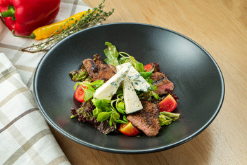 Grilled veal salad with lettuce, cherry tomatoes, dor blue cheese in a black bowl on wooden background. Selective focus, c;ose up