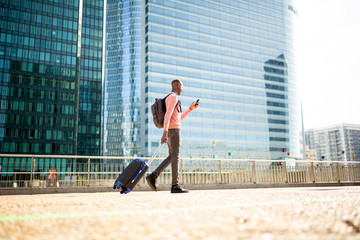 Fototapeta na wymiar Full body side young black man walking with phone and suitcase in city