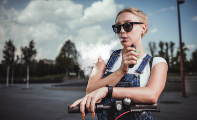 Plakat Modern girl smoking an e-cigarette as she is standing with electric scooter