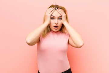 young pretty blonde woman feeling horrified and shocked, raising hands to head and panicking at a mistake against pink flat wall