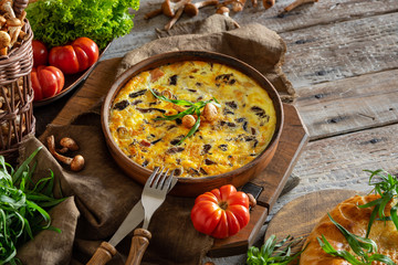 fritata or quiche with agaric mushrooms and cheese inceramic baking dish . Garnish with fresh tomatoes, lettuce and tarragon on a wooden table. Traditional Italian menu concept. Horizontal