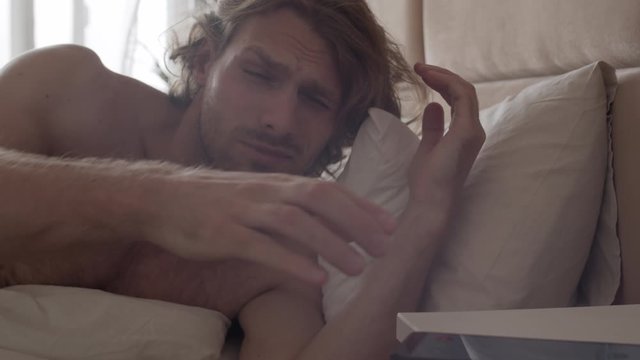 Slowmo shot of sleepy young man with stubble and long hair waking up in morning and checking time on his phone, then getting back to sleep