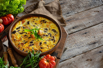 fritata or quiche with agaric mushrooms and cheese inceramic baking dish . Garnish with fresh tomatoes, lettuce and tarragon on a wooden table. Traditional Italian menu concept. Horizontal