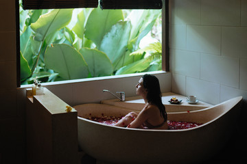 Sensitive young woman enjoying in spa. Luxury stone bath tub with jungle view near window. Natural organic tropical petals in the water. Beauty treatment concept.