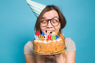 Close up funny positive girl in glasses and greeting paper hat holding a happy birthday cake in her...