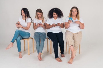 Fototapeta premium Group of beautiful girls sitting on chairs and using smartphones on white background. People and modern gadgets