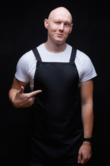 Strong handsome smiling bald guy wearing black apron pointing his finger on his chest. Butcher, baker, chef or waiter concept. Mockup for restaurants, grossery shops, bakery, butchery.