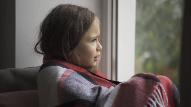 Close-up of a brunette girl covered with a warm blanket sitting on windowsill at home. Caucasian child with fever looking at the window sadly. Concept of health, illness, sickness, cold, treatment.