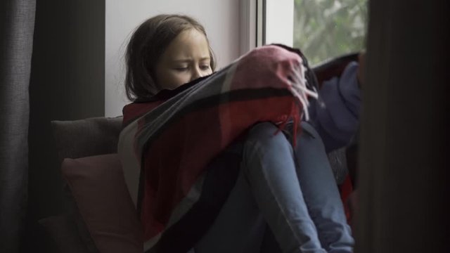 Sick brunette girl covered with a warm blanket sitting on windowsill at home. Caucasian child with fever looking at the window sadly. Concept of health, illness, sickness, cold, treatment.
