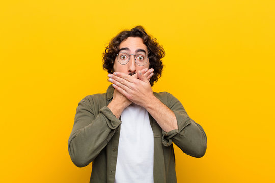 young crazy man covering mouth with hands with a shocked, surprised expression, keeping a secret or saying oops against yellow wall