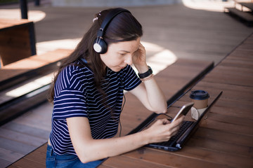 A girl student with a smartphone, sits at a laptop and listens carefully in headphones, learns...