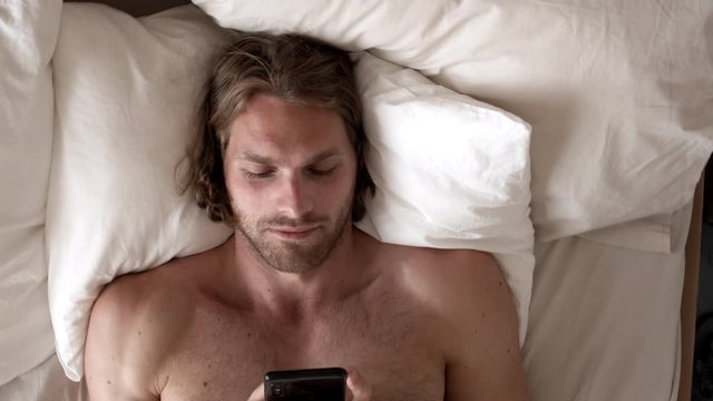 Top view rotating camera shot of handsome man with bare chest lying awake in bed in morning and typing on his mobile phone