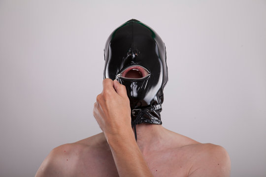 man wears black shiny latex mask on his head opening metal zipper over mouth
