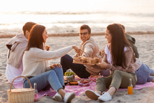friendship, leisure and fast food concept - group of happy friends eating sandwiches or burgers at picnic on beach in summer
