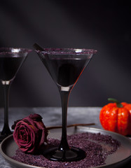 Two glasses with black cocktail, dried roses, pumpkin for Halloween party on the dark background