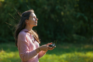 A woman in clearing in the park watches drone flying, looking into the distance, and holding control panel in her hands.