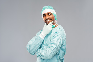 medicine, surgery and people concept - smiling thiniking indian male doctor or surgeon in protective wear over grey background