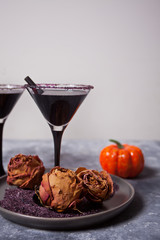 Two glasses with black cocktail, dried roses, pumpkin for Halloween party on the dark background