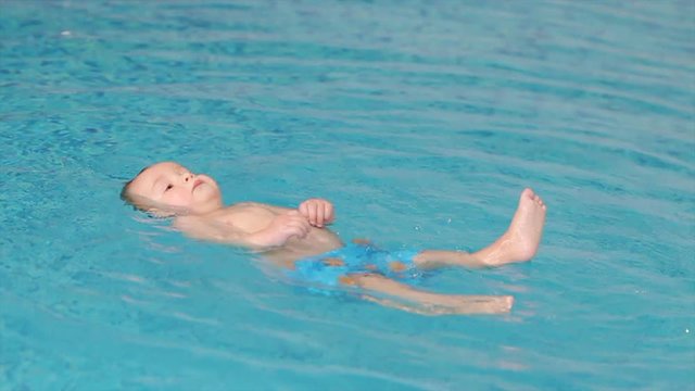 Cute toddler stays on the water in the swimming pool, swims on his back