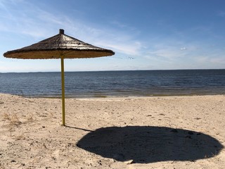 An umbrella stands on a sandy beach against the background of sky and water. Empty beach on the shore of a large reservoir. Shade from a solar umbrella.