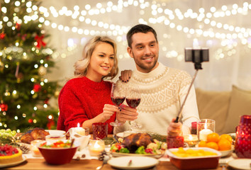 christmas, holidays, technology and people concept - happy couple in taking picture by selfie stick at home dinner