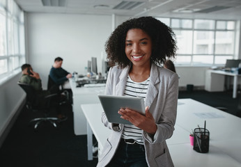 Portrait of a smiling confident african american young businesswoman holding digital tablet in hand looking at camera with colleague at background in office