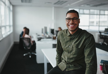 Portrait of a successful smiling young professional businessman sitting over desk in front of colleague working in background - Powered by Adobe