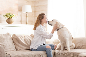 Young pretty woman in casual clothes hugging her beloved big white dog sitting on the sofa in the...