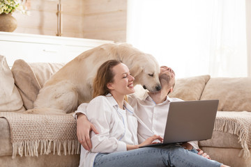 Young happy positive married couple charming woman and young man all sit on the floor next to their beloved dog and watch cheap airline tickets for summer holidays on the Internet using a laptop