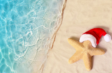 Fototapeta na wymiar Christmas holidays concept. Top view of starfish in santa claus hat on summer sand beach.