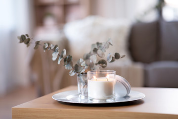 decoration, hygge and cosiness concept - burning white fragrance candle on tray and branches of eucalyptus populus on table at cozy home