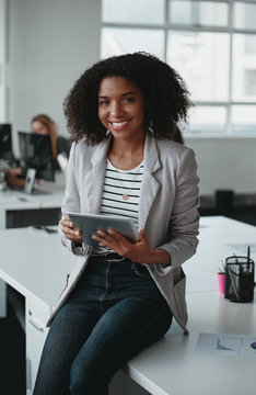 Happy confident african american business woman employee holding digital tablet looking at camera sitting on desk in office