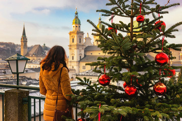 Young woman view from the back stands on the background of sunny Christmas Salzburg, Austria. Christmas trees with red Christmas balls against the background of the winter Salzburg. - obrazy, fototapety, plakaty