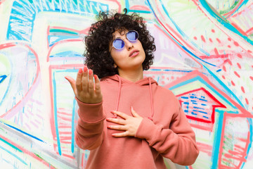 young pretty afro woman feeling happy and in love, smiling with one hand next to heart and the other stretched up front against graffiti wall